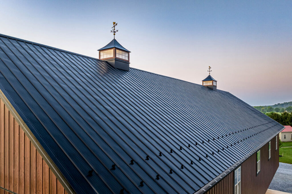 Metal corrugated roof with two cupolas and weather vanes, viewed from above at sunset over a wooden building side.
