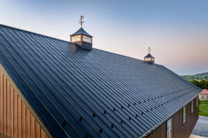 Metal corrugated roof with two cupolas and weather vanes, viewed from above at sunset over a wooden building side.