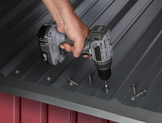Person using a cordless drill to drive a screw into a metal roof panel with several screws nearby.
