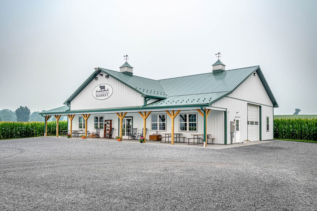 White market building with a green metal roof and a long covered porch, outdoor chairs and planters along a gravel lot, cornfields in the background, sign reads 'Beaver Creek Farms Market'