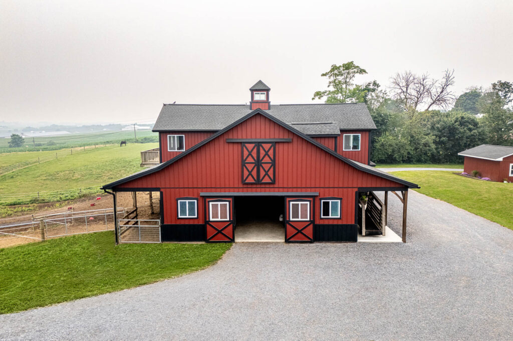 Red barn with black trim and cross-buck doors, set in a rural farmyard with a gravel driveway and green fields beyond.