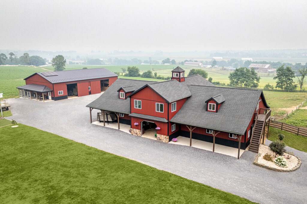 A red barn-style farmhouse with a gray roof, cupola, and covered porches on a gravel driveway amid green fields.